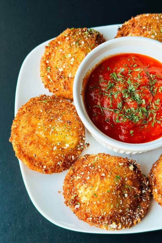 Fried ravioli on a white plate with a small bowl of sauce